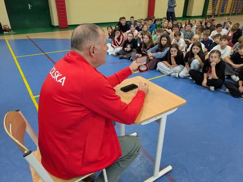 Spotkanie z Olimpijczykiem Marianem Woroninem na sali gimnastycznej. Olimpijczyk prezentuje medal olimpijski i opowiada o swojej karierze sportowej. Spotkanie odbywa się na sali gimnastycznej Szkoły Podstawowej nr 6