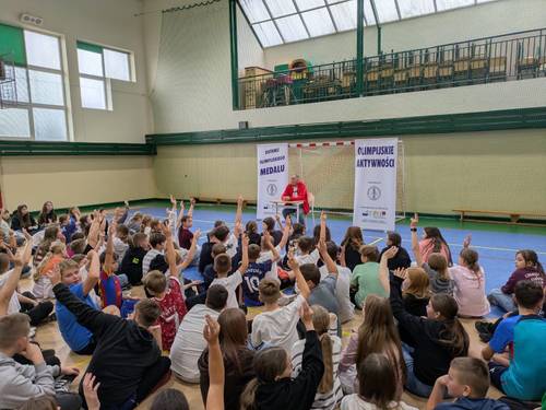 Spotkanie z Olimpijczykiem Marianem Woroninem na sali gimnastycznej. Olimpijczyk prezentuje medal olimpijski i opowiada o swojej karierze sportowej. Spotkanie odbywa się na sali gimnastycznej Szkoły Podstawowej nr 6