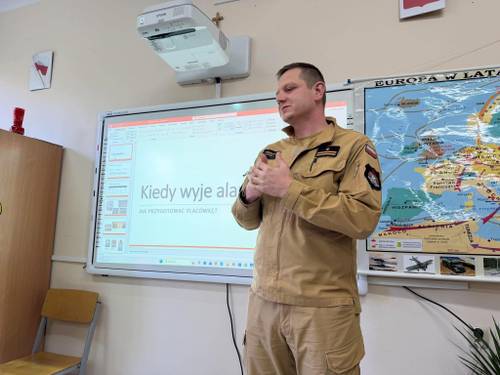 Funkcjonariusz Strazy Pożarnej omawia organizacje i przebieg ćwiczeń. W tle elektroniczna tablica edukacyjna z rodzajami alarmów